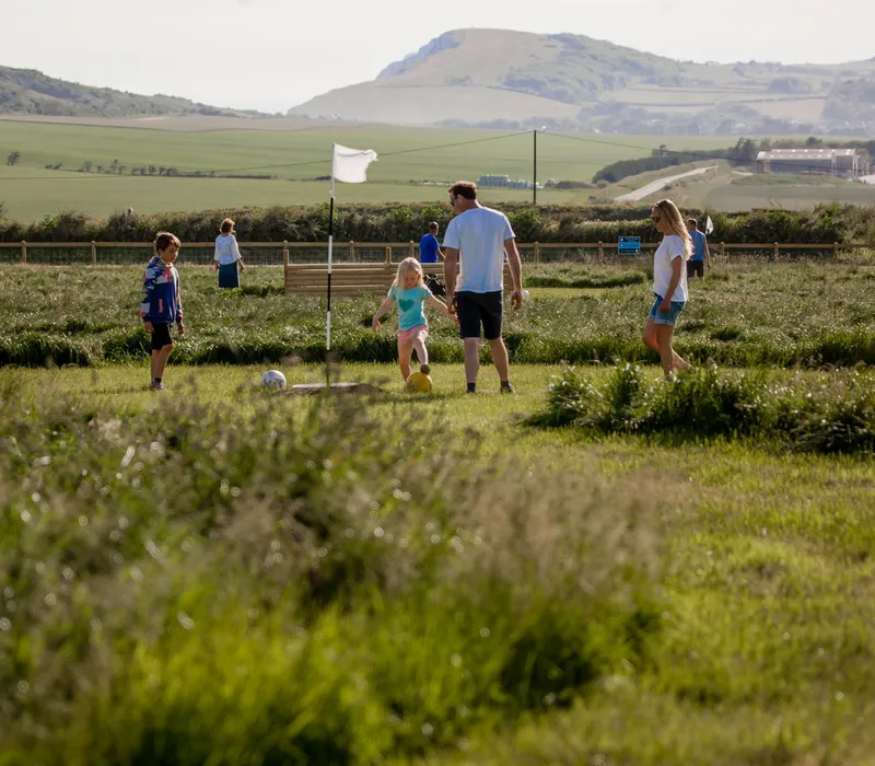 Family playing football golf on a grassy course at Tapnell Farm with a flag marker and countryside hills in the background