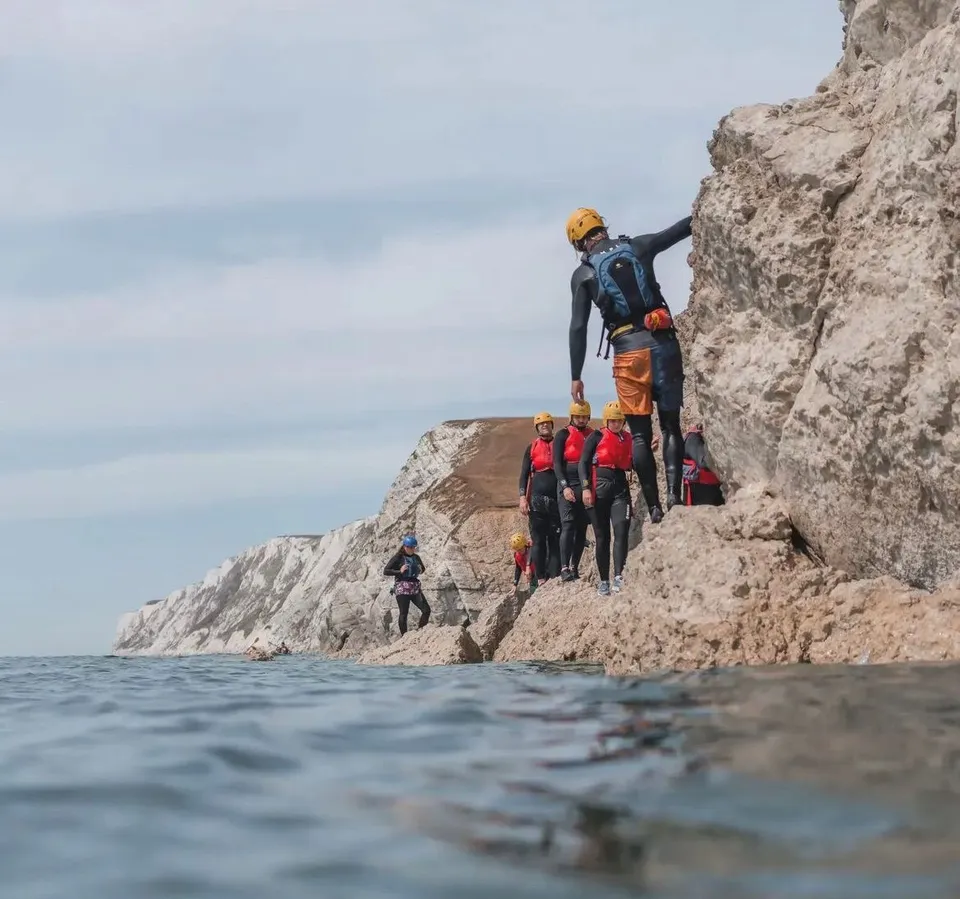 Group coasteering along a rocky cliff at Freshwater Bay, wearing helmets and buoyancy aids beside the sea