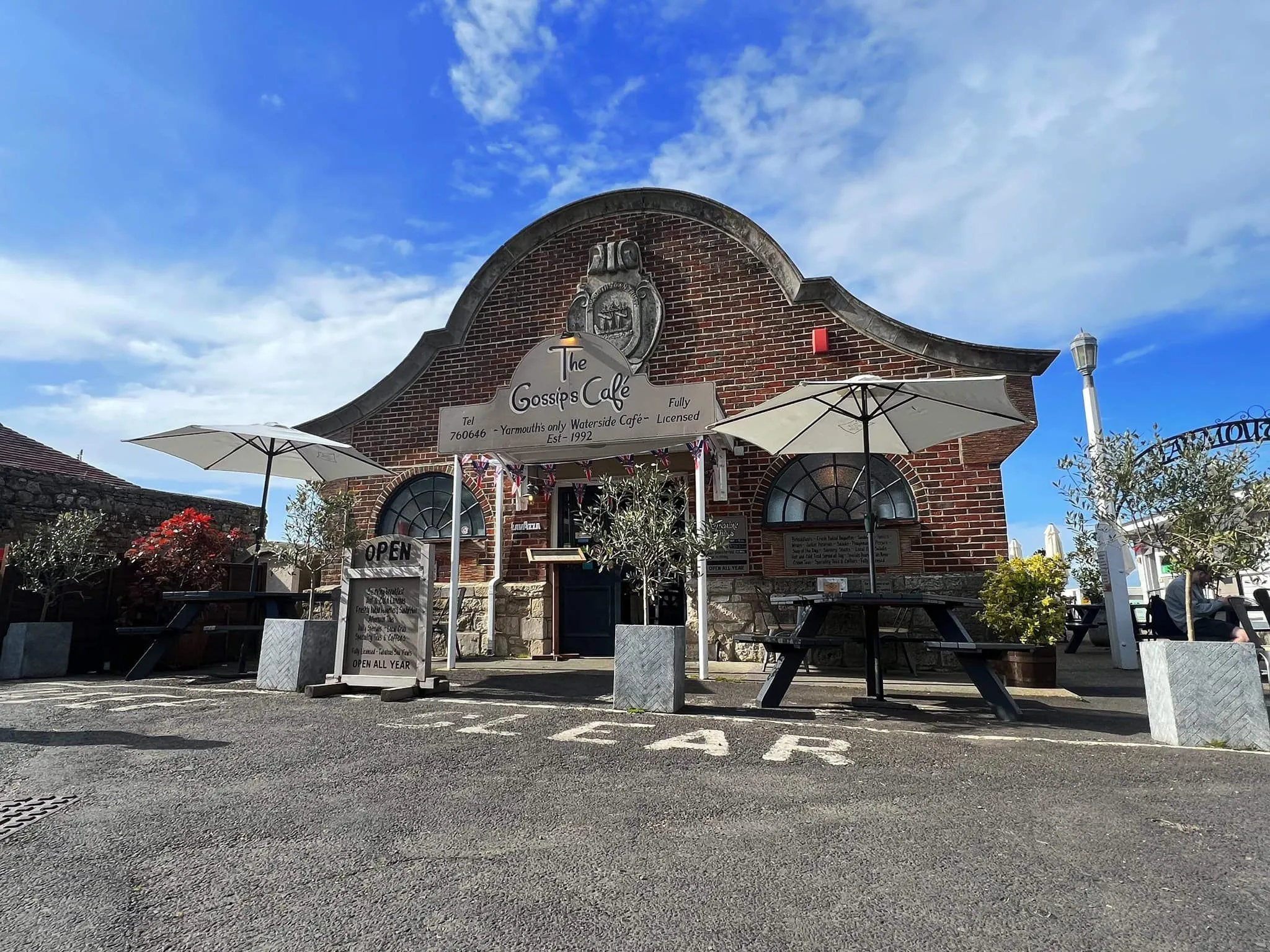 Exterior of The Gossips Café, a brick waterside café with outdoor tables and parasols under a blue sky