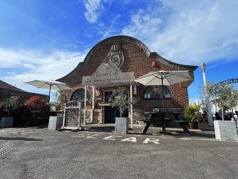 Exterior of The Gossips Café, a brick waterside café with outdoor tables and parasols under a blue sky