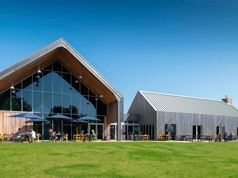 Modern barn-style building with large glass front and outdoor seating on a lawn under a clear blue sky