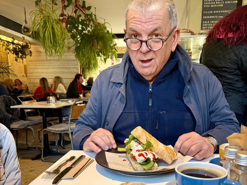 Man wearing glasses sitting in a café with a baguette sandwich and coffee on the table