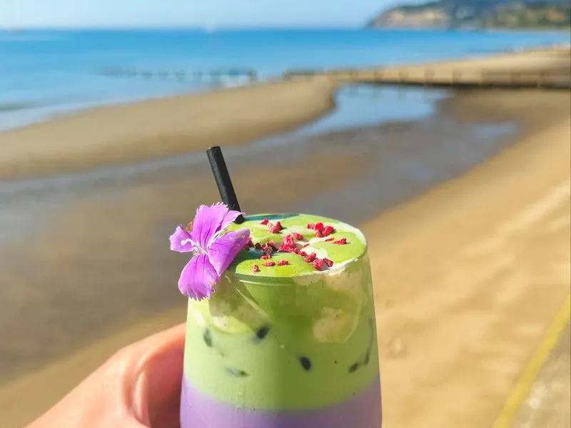 Hand holding a layered purple and green iced drink with a flower garnish on a sandy beach