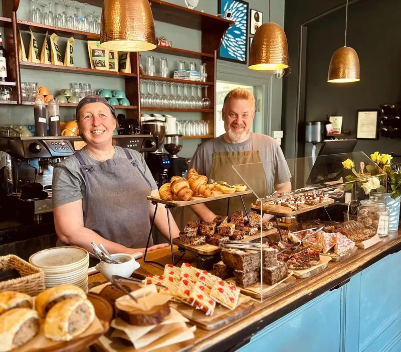 Two café staff smiling behind a counter filled with pastries, cakes and bread under warm copper pendant lights.