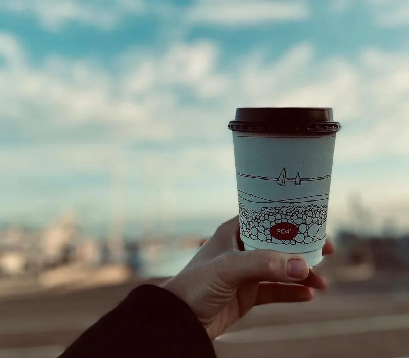 Hand holding a takeaway coffee cup with an illustrated seaside design against a cloudy sky