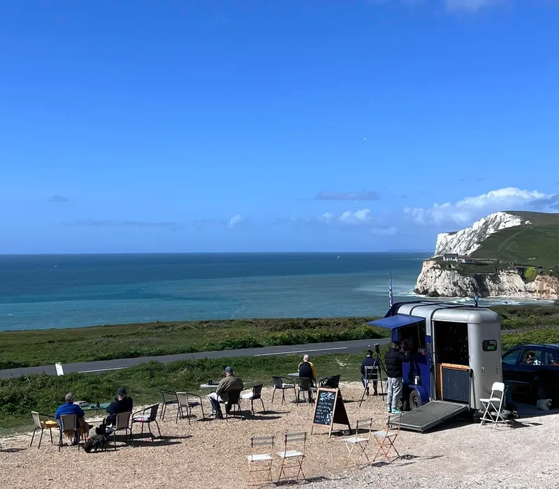 Small coffee trailer with outdoor seating overlooking the sea and white cliffs under a clear blue sky.