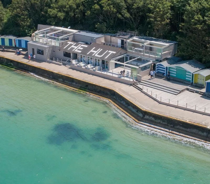 Aerial view of The Hut beachfront building with colourful beach huts beside turquoise sea