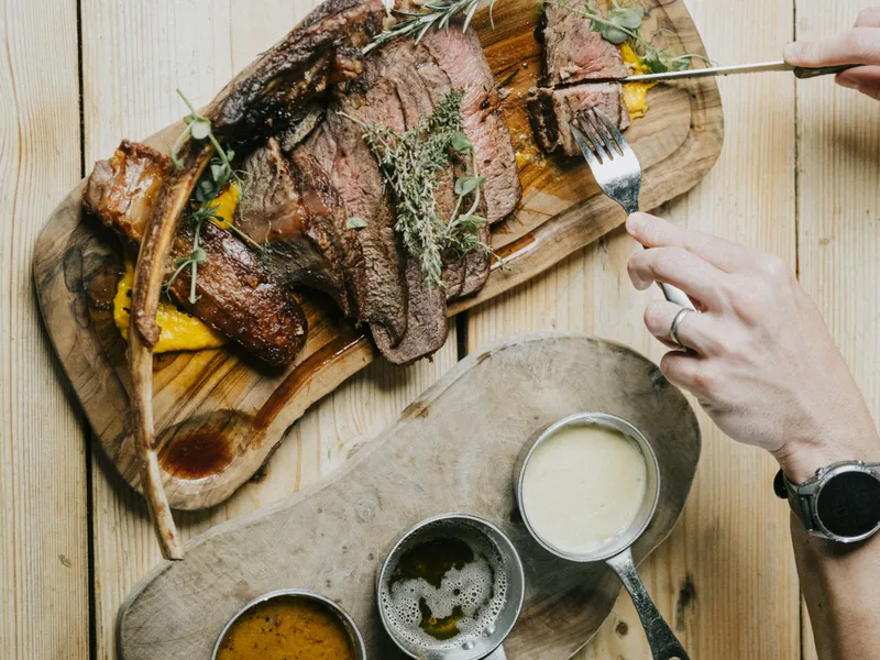 Sliced tomahawk steak on a wooden board with herbs, being cut with a knife and fork