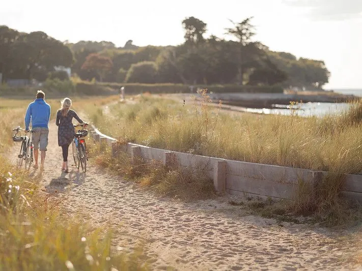 Two people walk barefoot along a sandy path pushing bicycles beside coastal dunes at sunset