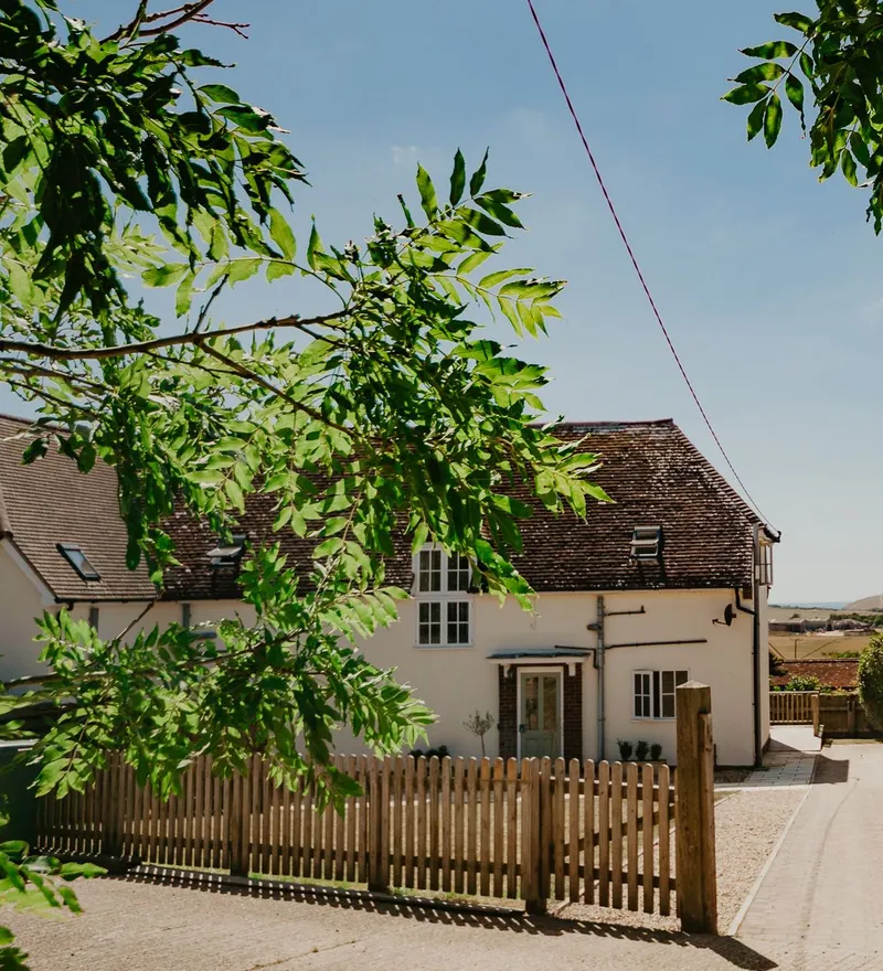 White countryside cottage with a wooden picket fence and leafy branches under a blue sky
