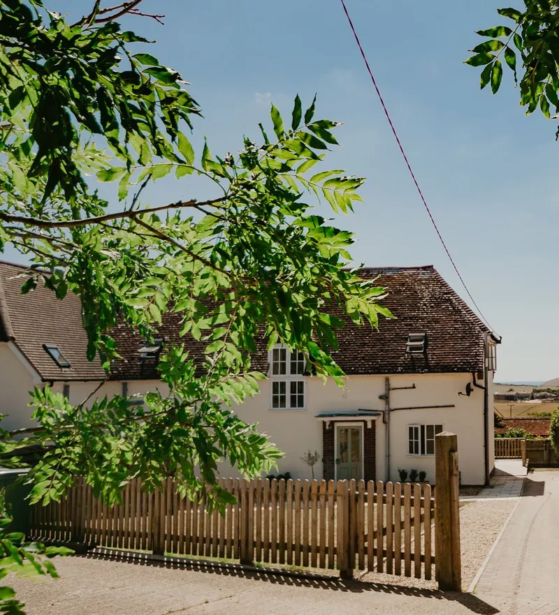 White countryside cottage with a wooden picket fence and leafy branches under a blue sky