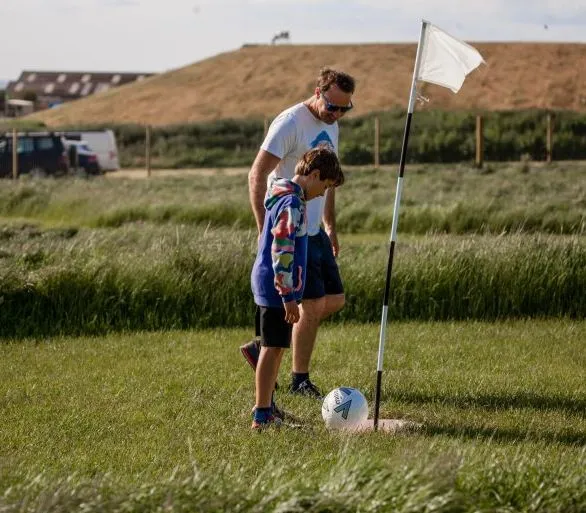 Man and child playing football golf, standing next to a flag on a grassy field.