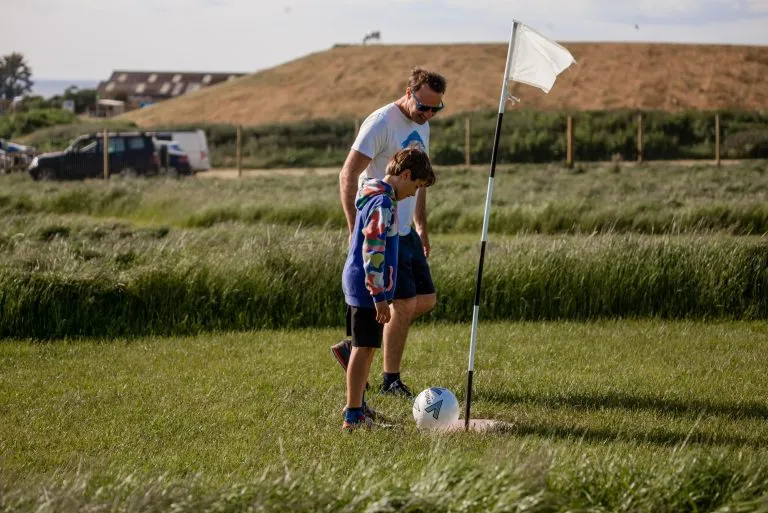 Man and child playing football golf, standing next to a flag on a grassy field.