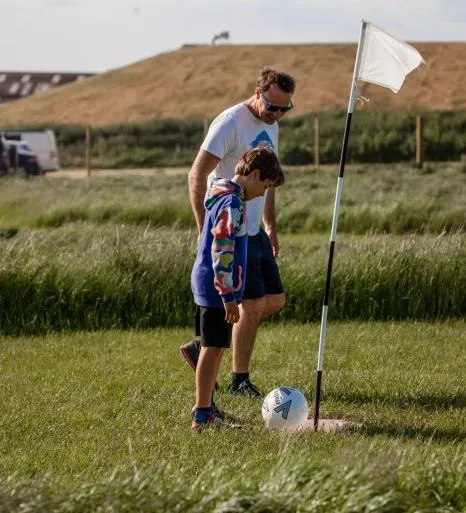Man and child playing football golf, standing next to a flag on a grassy field.