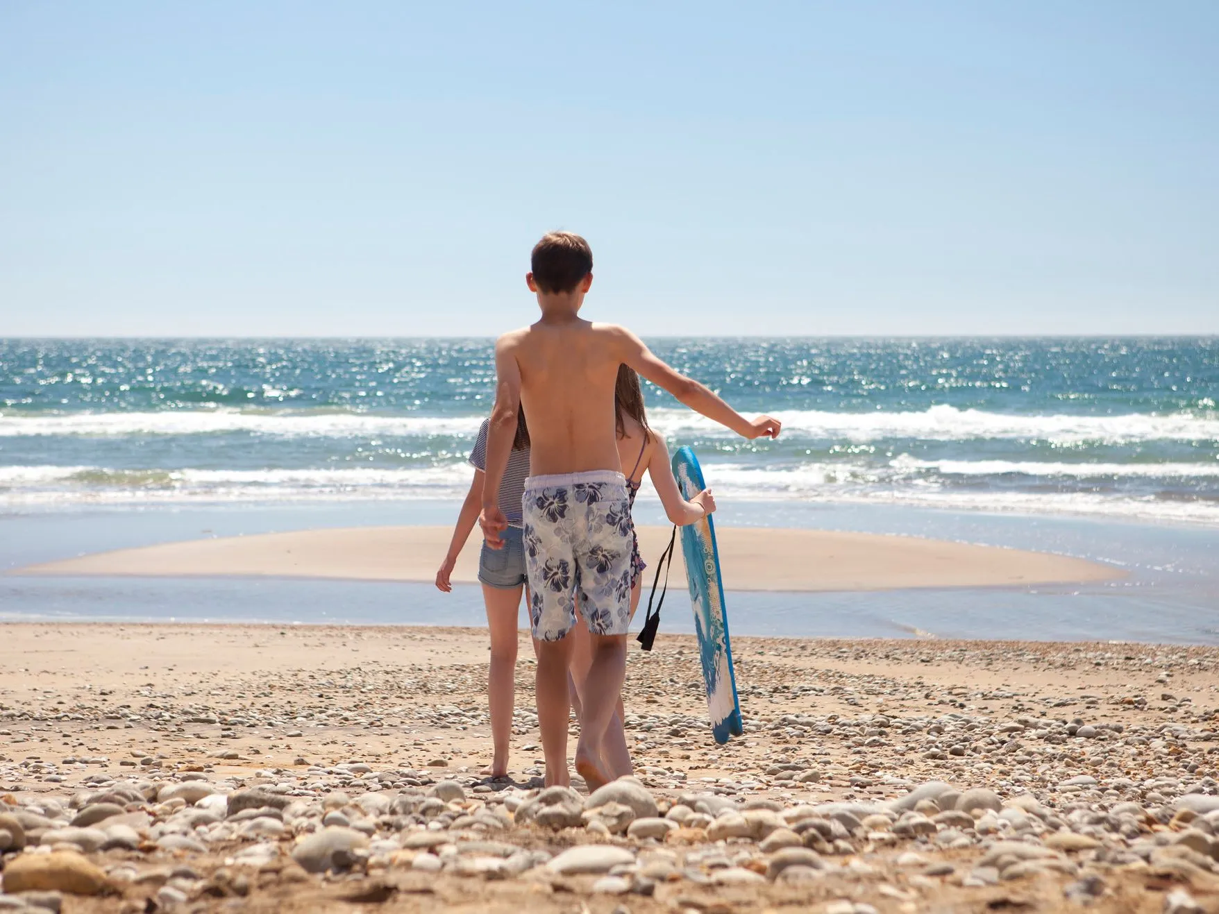 Three children walk towards the sea on a pebbly beach, one carrying a blue skimboard.