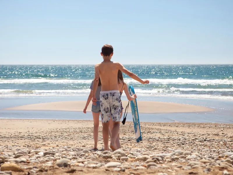 Three children walk towards the sea on a pebbly beach, one carrying a blue skimboard.