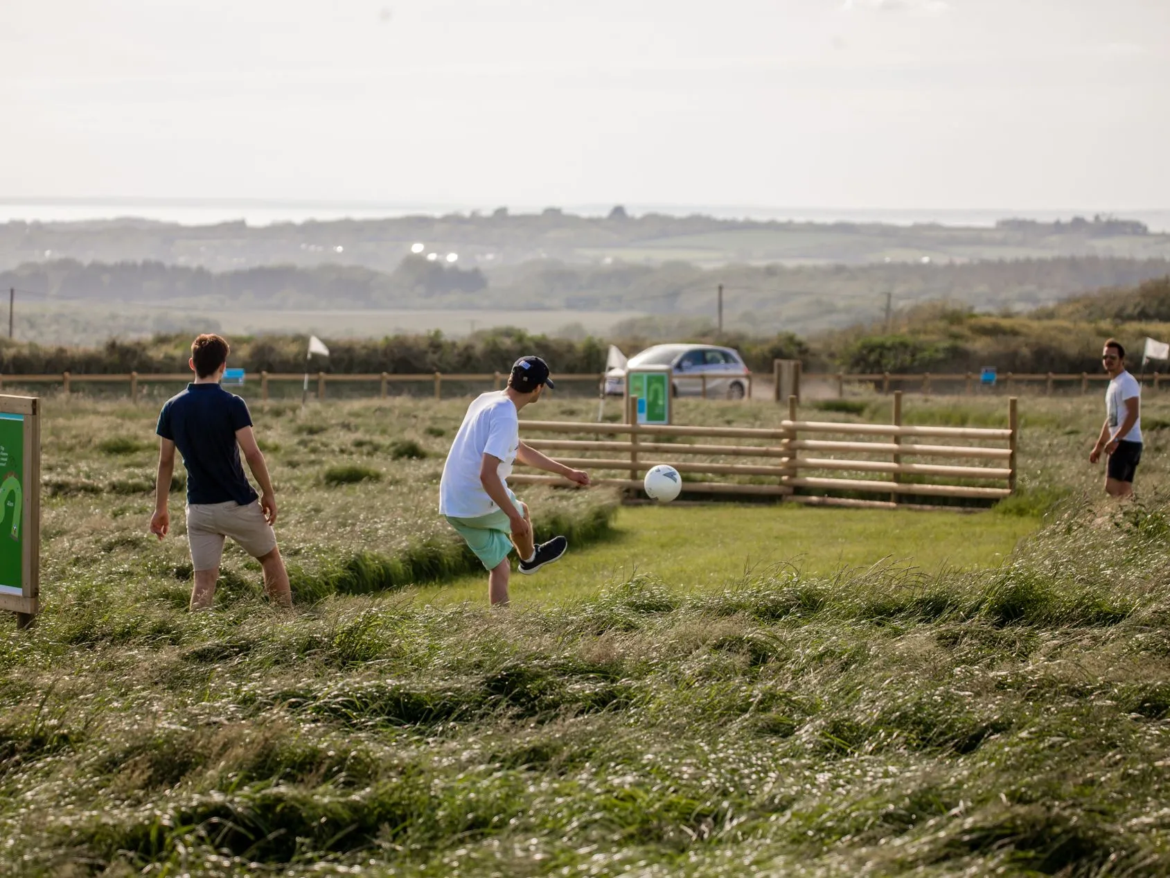 Three friends playing football golf on a grassy course at Tapnell Farm, with one kicking a ball near a hole sign