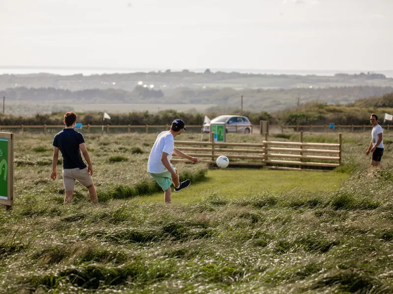 Three friends playing football golf on a grassy course at Tapnell Farm, with one kicking a ball near a hole sign