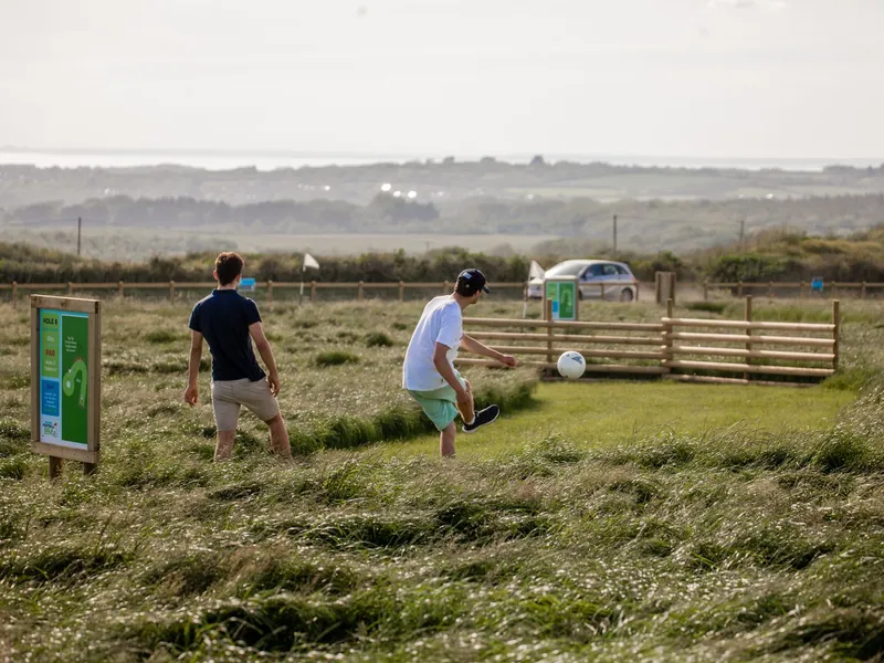 Three friends playing football golf on a grassy course at Tapnell Farm, with one kicking a ball near a hole sign