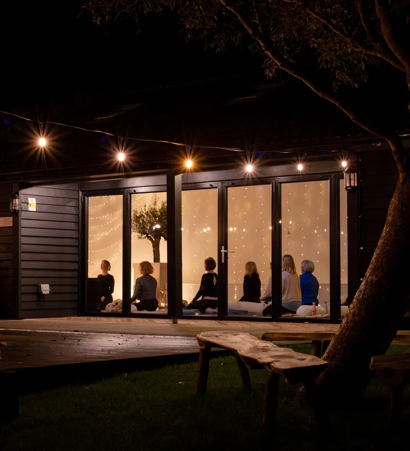 Evening view of a cosy studio lit with fairy lights, with a small group seated on yoga mats inside.