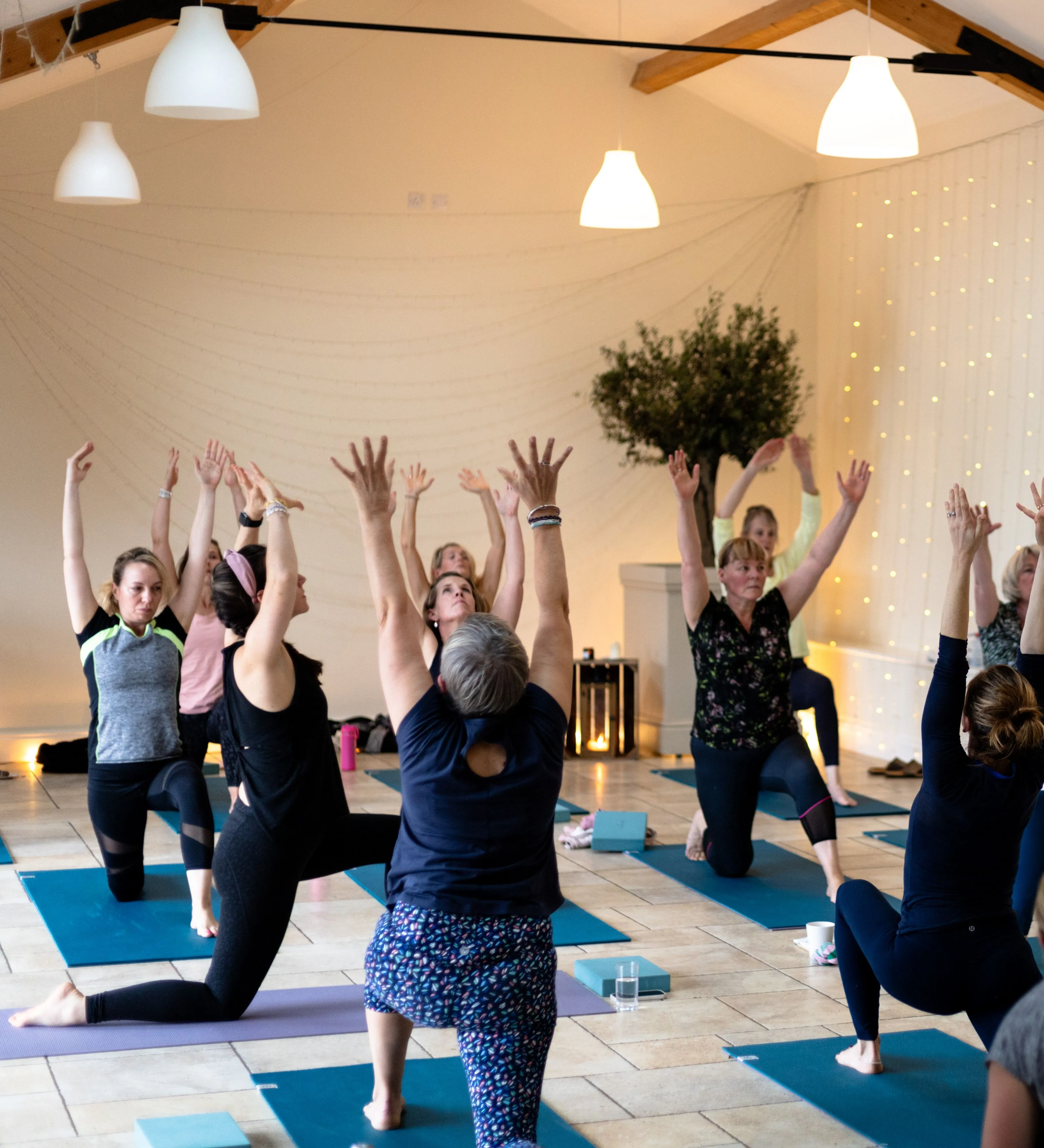 Group yoga class with participants raising their arms in a bright studio lit by fairy lights