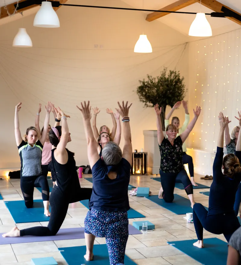 Group yoga class with participants raising their arms in a bright studio lit by fairy lights