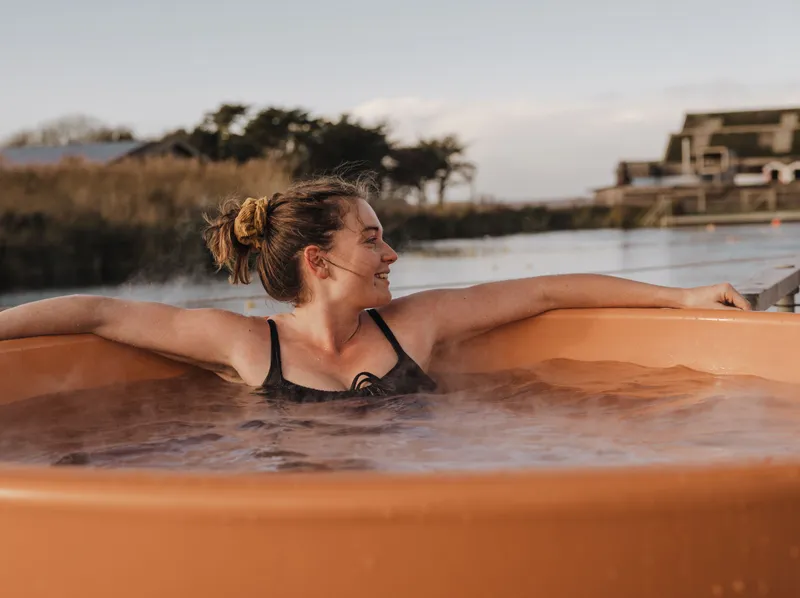 Woman relaxing in an outdoor hot tub by a waterside at sunset