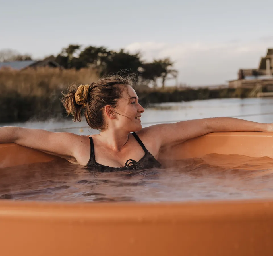 Woman relaxing in an outdoor hot tub by a waterside at sunset