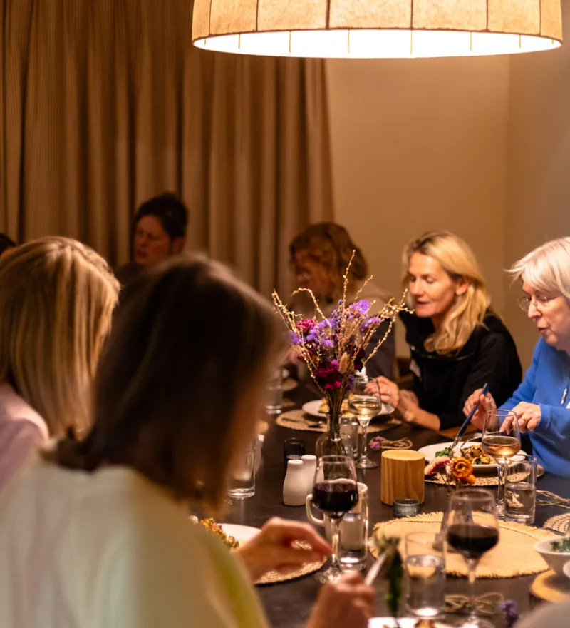 Guests enjoying a meal together at a long dining table under a large warm pendant light.