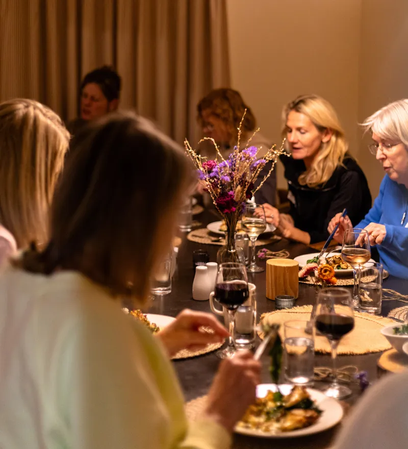 Guests enjoying a meal together at a long dining table under a large warm pendant light.
