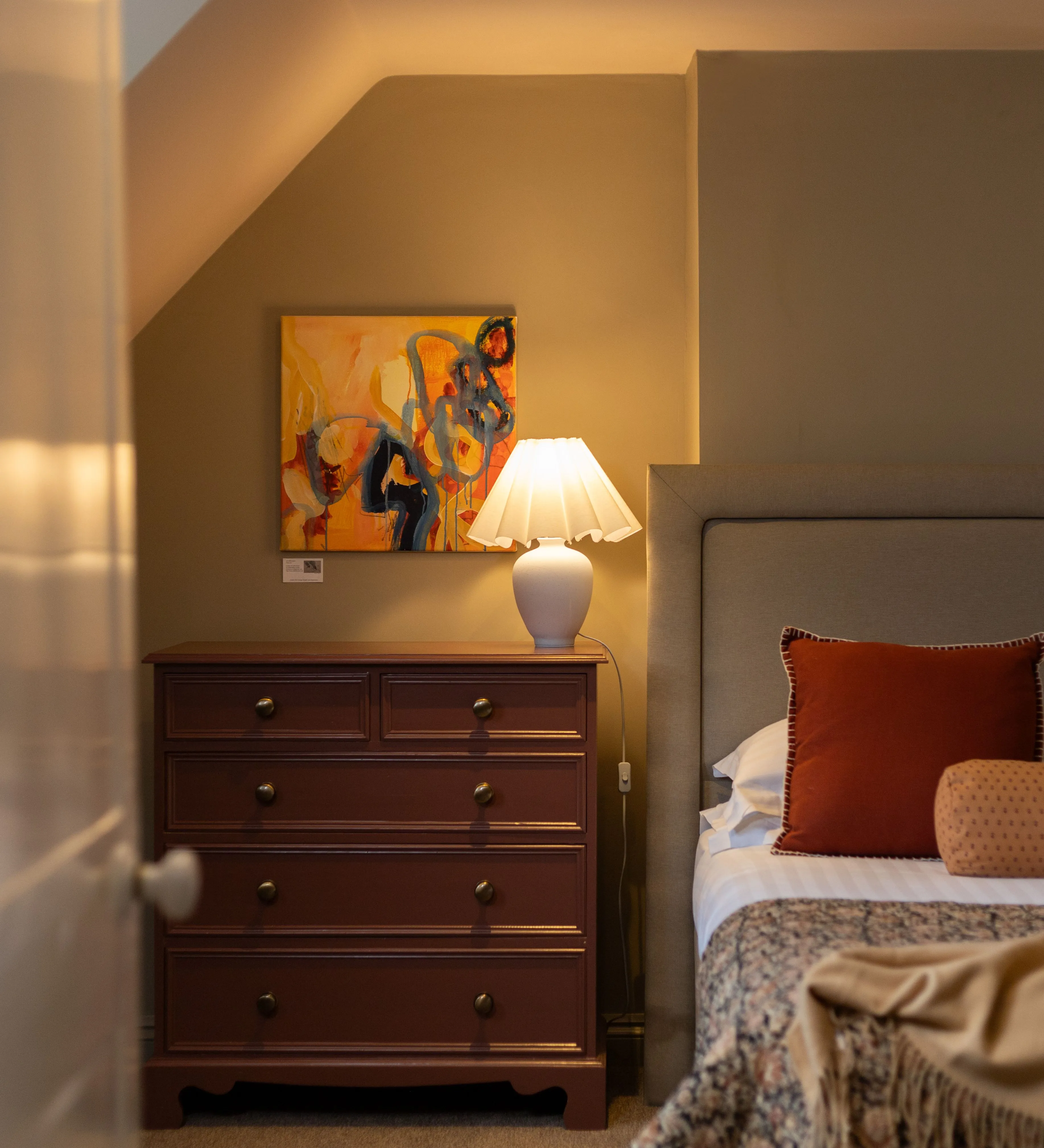 Cosy bedroom interior with upholstered headboard, bedside chest of drawers, lamp and colourful artwork