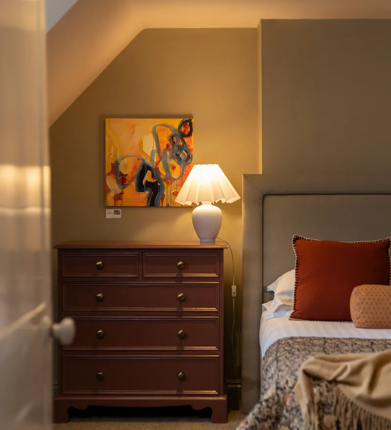 Cosy bedroom interior with upholstered headboard, bedside chest of drawers, lamp and colourful artwork