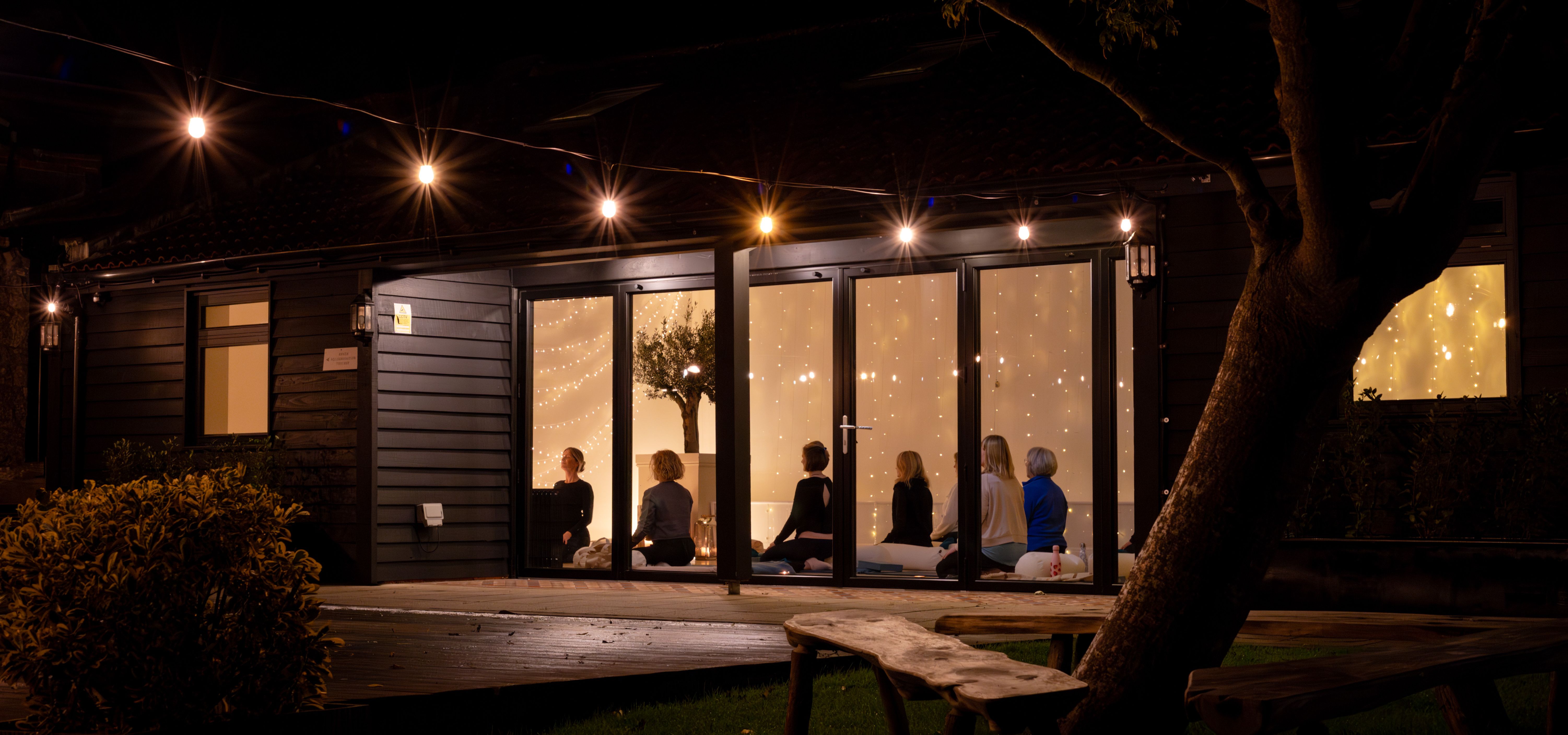 Night-time yoga class inside a warmly lit studio with fairy lights and outdoor string lights