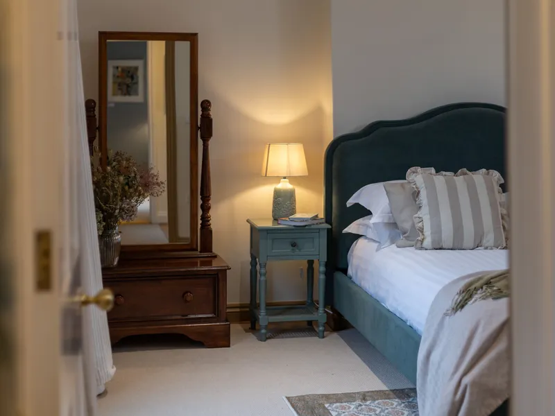 Cosy bedroom with teal upholstered bed, bedside table and lamp, and wooden dresser with mirror