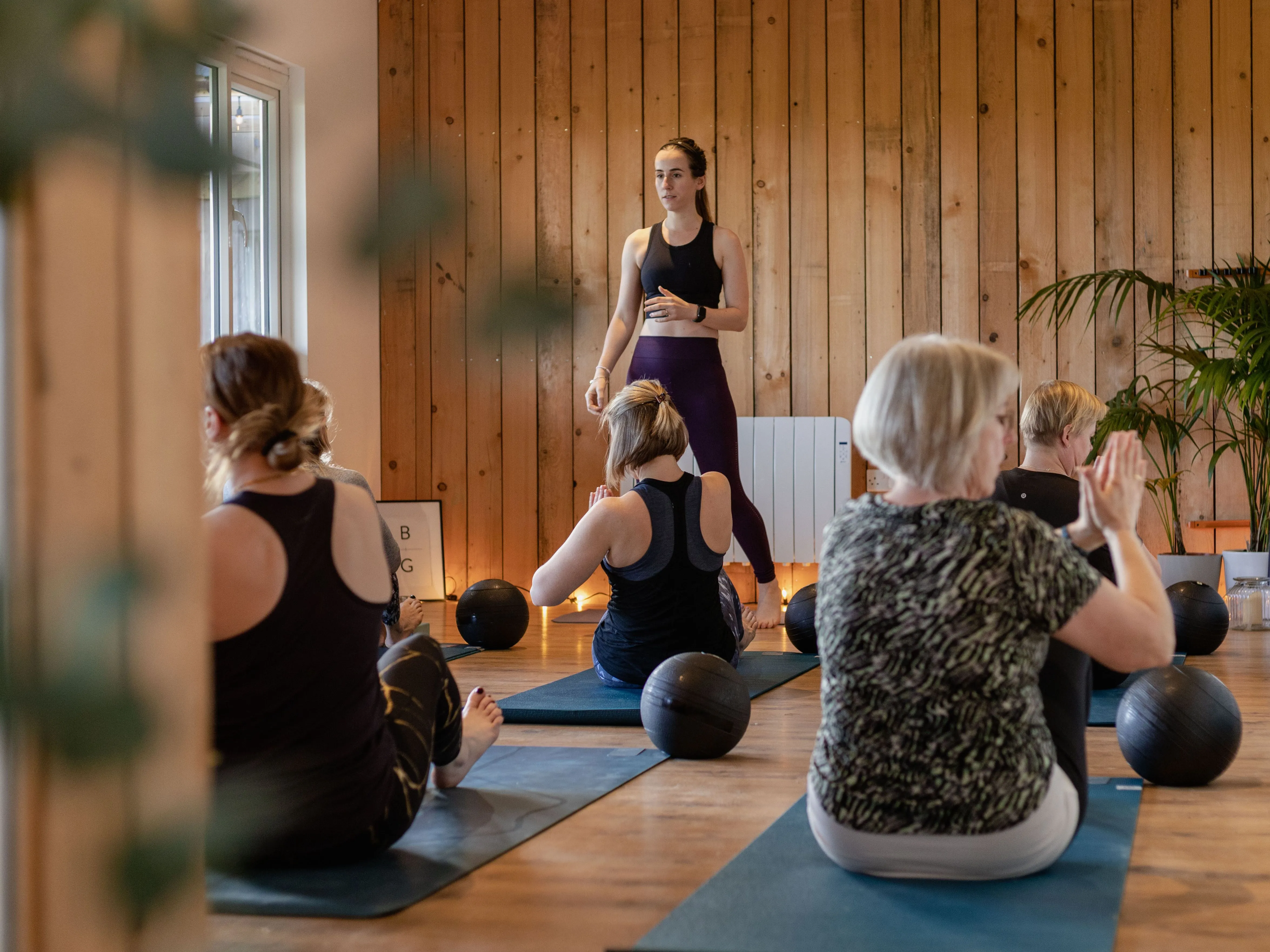 Yoga instructor leading a class in a wooden studio with participants seated on mats