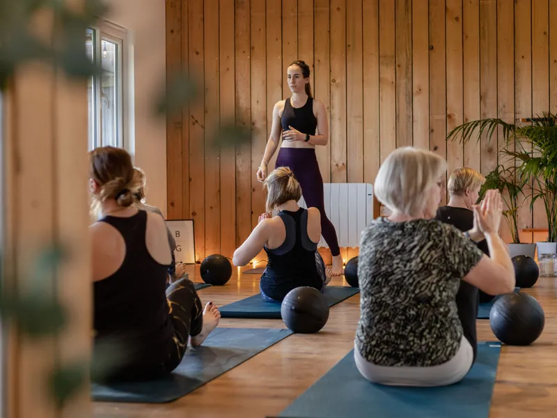 Yoga instructor leading a class in a wooden studio with participants seated on mats