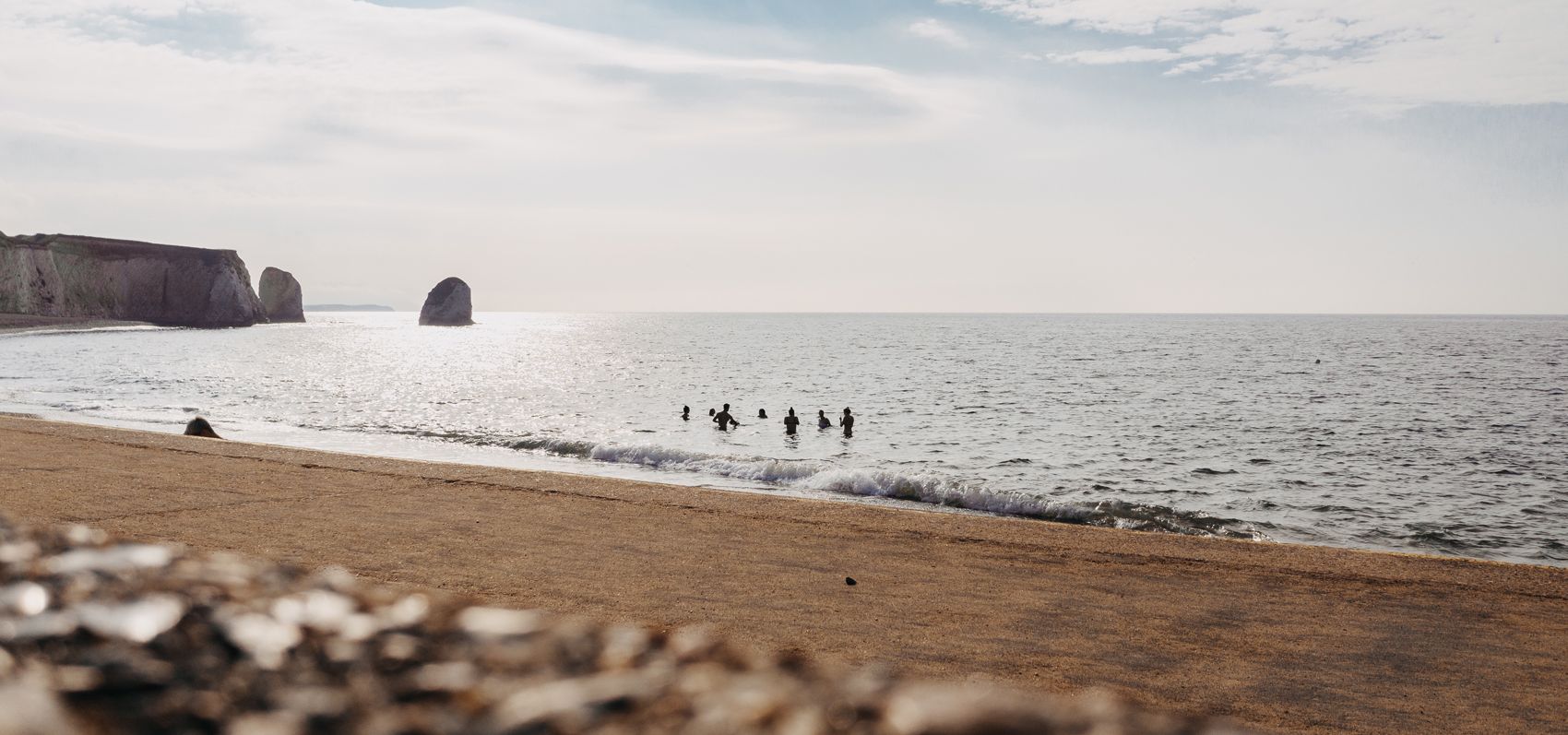 A group of swimmers in the sea near a sandy beach with cliffs and rock stacks in the distance