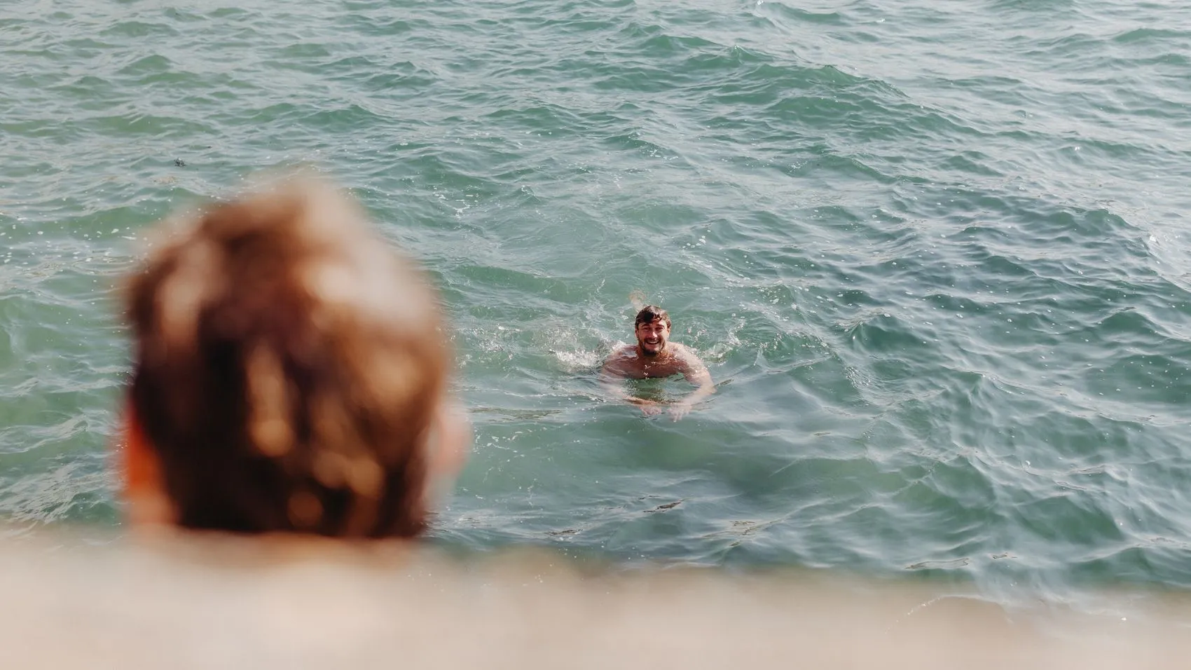 Smiling man swimming in the sea, viewed from shore with a blurred person in the foreground