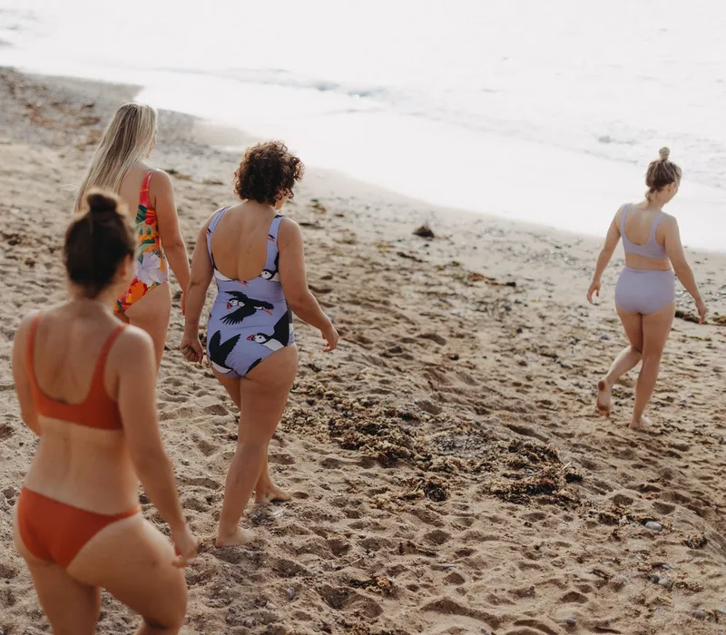 Four women in swimsuits walking along a sandy beach towards the sea