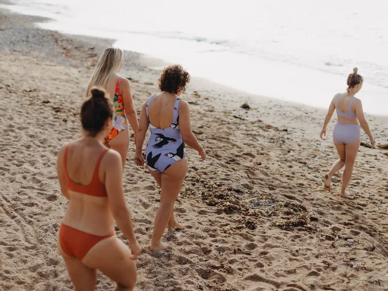 Four women in swimsuits walking along a sandy beach towards the sea