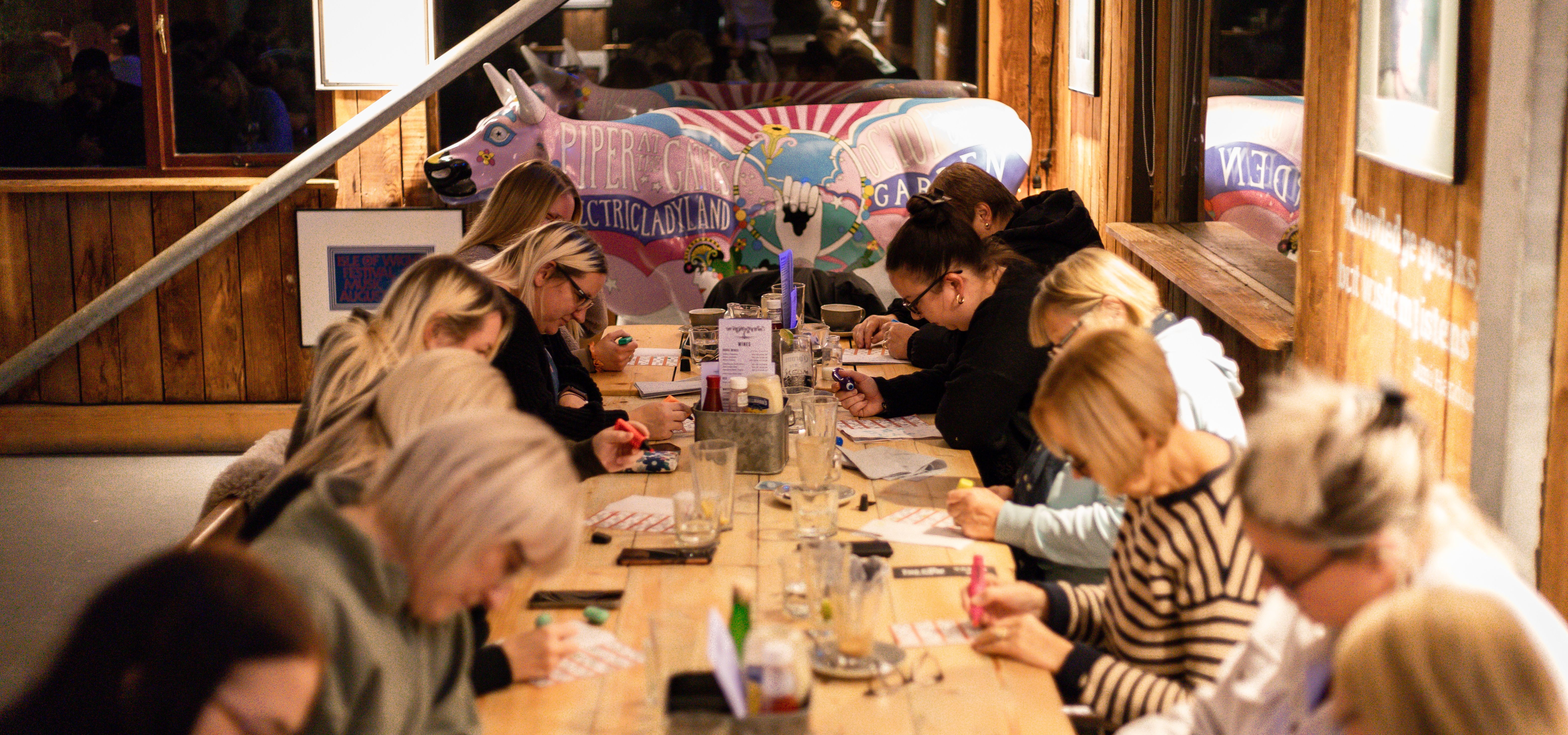 Guests fill in bingo cards at a long table during Tapnell Farm bingo night, with a colourful cow statue in the background.