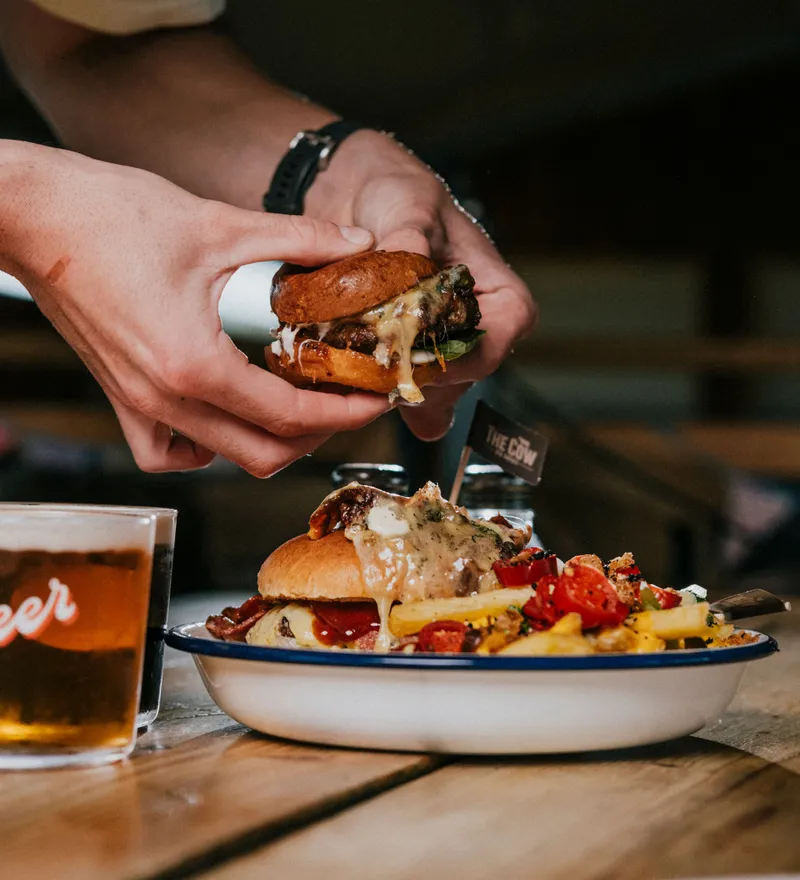 Hands holding a cheeseburger above a plate with a burger, chips and salad on a wooden table, with a pint of beer nearby