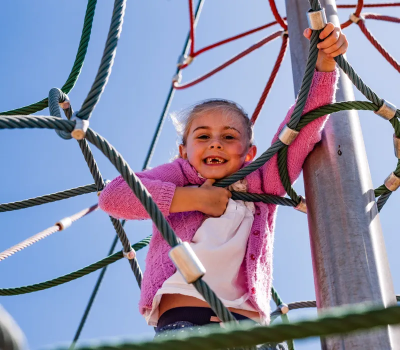Smiling child climbing a rope web play frame under a clear blue sky