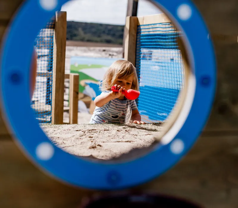 Toddler playing with sand using a red scoop, framed through a blue circular tunnel on a playground
