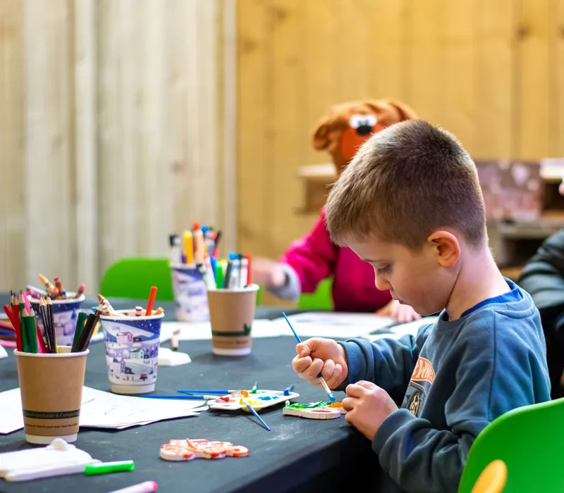 Child painting at a craft table with pencils, paper and paints while adults and a toddler sit nearby