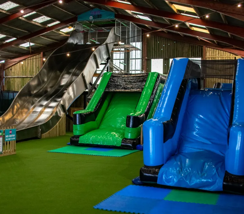 Indoor play area with a large metal slide and green and blue inflatable slides under a barn roof