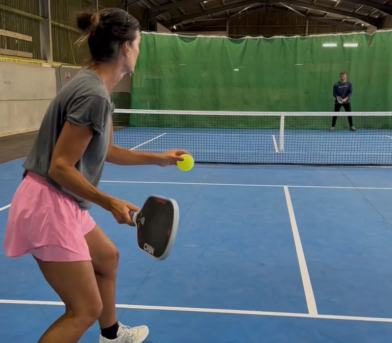 Player serving a pickleball on an indoor court with an opponent waiting across the net