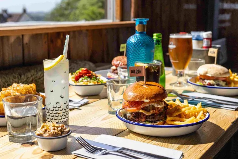Table set with burgers, fries, salads, drinks, and condiments in a sunny restaurant setting.
