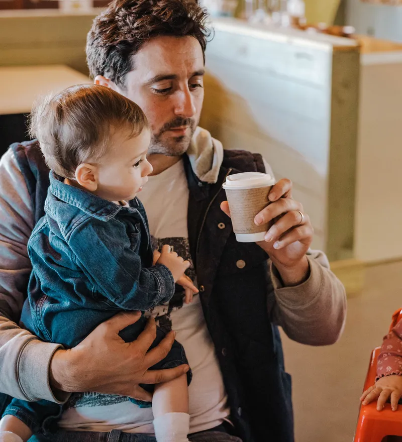 Adult holding a toddler and a takeaway coffee while another toddler sits in a high chair indoors at Tapnell Farm Park