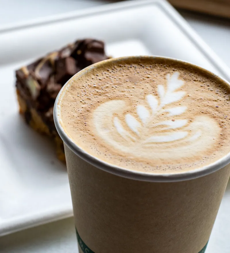 Pumpkin spiced latte with leaf latte art in a takeaway cup on a tray, with a chocolate brownie in the background